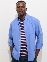 A man with short hair and a neutral expression stands against a white background, wearing the Mill Shirt in Cotton Poplin—a light blue 100% cotton button-down—over a navy tee with red, white, and blue stripes, hands in his pockets.