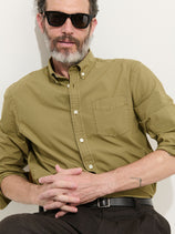 A man with a salt-and-pepper beard, in black sunglasses and the Mill Shirt In Cotton Poplin in olive green, sits against a white wall with his hands clasped on his lap.