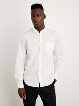 A man stands against a plain white background, wearing the Mill Shirt in Cotton Poplin with French seams and real shell buttons, paired with black pants and one hand in his pocket.