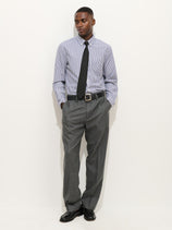 A man stands against a white background, wearing the Mill Shirt In Stripe Portuguese Poplin with a black tie, gray dress pants, and black shoes. His hands are in his pockets as he looks slightly to the side.