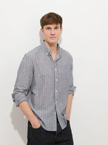 A young man with short brown hair stands against a plain white background, wearing the Mill Striped Shirt In Chambray with sleeves rolled up and dark pants, hands in his pockets, looking at the camera with a neutral expression.