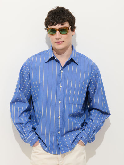 A man wearing the Franklin Striped Shirt in cotton poplin, featuring blue and white vertical stripes and real shell buttons, pairs it with green sunglasses while standing against a plain white background with hands in pockets.