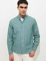 A man stands against a plain white background wearing the Mill Shirt In Thin Stripe—a green and white vertically striped button-down over a light blue t-shirt—showcasing effortless style.