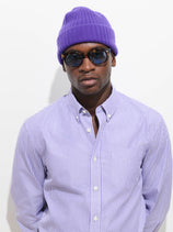 A man wearing the Cashmere Beanie in purple, paired with a striped purple button-up shirt and sunglasses, stands against a white background.