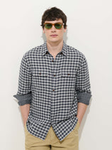 A man wearing sunglasses, the Mill Double Gauze Shirt in Cotton Plaid (black and white) with rolled-up sleeves, and beige pants stands against a plain white background with his hands in his pockets.