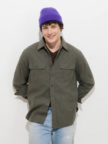 A person wearing a purple beanie, dark shirt, the Potter Shirt in Chamois (a gray 100% cotton overshirt), and light blue jeans stands against a white background, smiling with hands behind their back.