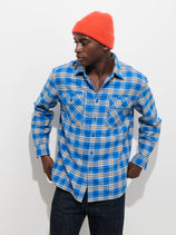 Wearing the Cyrus Shirt Flannel in blue plaid, paired with a bright orange beanie and dark jeans, a person stands against a white background, looking to the side with their hands in their pockets.