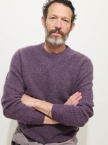 A man with curly dark hair and a salt-and-pepper beard stands against a white background, wearing the Jordan Sweater In Washed Cashmere in purple, arms crossed and showing a neutral expression.