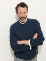 A man with curly dark hair and a beard stands against a white background, wearing the Donegal Crewneck Sweater in dark blue over a white collared shirt. He crosses his arms and looks confidently at the camera.