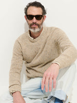 A man with a salt-and-pepper beard, dark sunglasses, and light blue jeans wears a beige Donegal Crewneck Sweater while sitting against a white background, one knee raised and his arm resting on it.