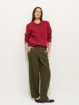 A woman stands against a white background wearing a red sweater, black flat shoes, and the 9th Street Pant—pigment-dyed cotton women’s artist pants with wide legs. Her hands are in her pockets and her expression is neutral.