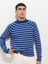 A young man with short dark hair wears the Alex Striped Rollneck In Cotton—a blue and white striped 100% cotton sweater—paired with white pants. He stands against a plain white background, slightly lifting the sweater’s hem with one hand.
