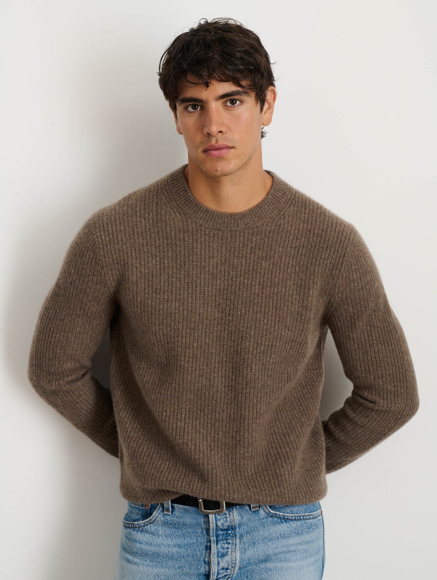 A young man with short dark hair wears the Jordan Sweater in Washed Cashmere and light blue jeans, standing against a plain white background with his hands behind his back, looking directly at the camera.