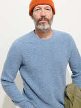 A man with a gray beard wears the Jordan Sweater In Washed Cashmere in light blue and an orange beanie, posing against a plain background and holding a green jacket in his left hand.
