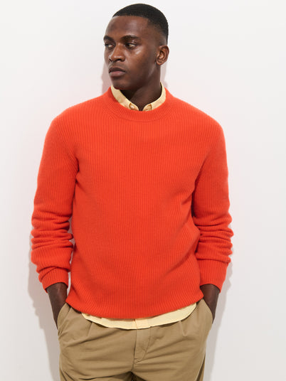 A man wears the Jordan Sweater in lightweight cashmere, bright orange, over a yellow collared shirt and khaki pants, standing against a white background with hands in his pockets, looking to the side.