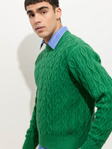 A young man with short dark hair wears the Aaron Cable Sweater in Cotton, styled over a blue collared shirt, standing against a plain light background and looking slightly to the side.