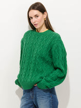 A woman with long brown hair wears the Aaron Cable Sweater in Cotton, a bright green relaxed-fit knit, paired with blue jeans. She stands against a plain white background with her hands in her pockets, looking at the camera.