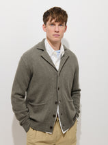 A young man with short brown hair stands against a plain white background, hands in his pockets, wearing the Alain Blazer in Cotton Linen over a white collared shirt and beige pants.