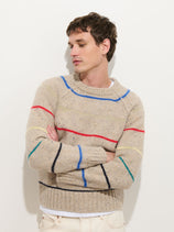 A person with short brown hair stands with arms crossed, wearing the Freddie Raglan Striped Crewneck Sweater in Merino Wool—a beige knit sweater featuring colorful stripes—while looking to the side against a plain white background.