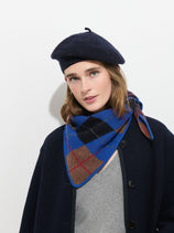 A person wearing a dark beret, a gray top, a dark coat, and the Flynn Argyle Handkerchief Scarf in Wool stands against a white background, looking at the camera.