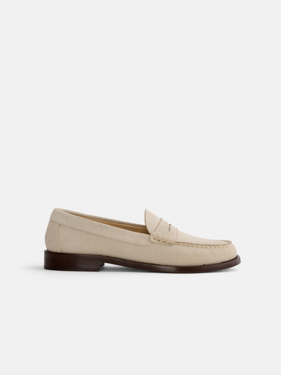 The New York Loafer Sister In Suede is a beige handsewn suede loafer featuring visible stitching, a penny slot detail, and a low brown heel, shown in profile on a plain white background.