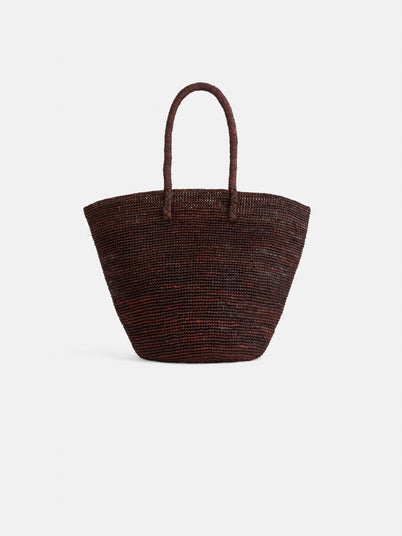 The Mary Mill Tote is a dark brown handwoven raffia bag with two rounded handles and a wide, rounded trapezoid shape. Its textured surface makes it a chic summer accessory, shown here against a plain white background.