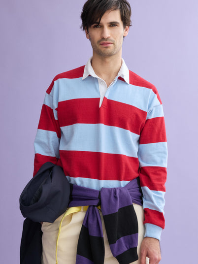 A man with dark hair wears a red and light blue J.Press x Alex Mill Rugby Striped Shirt in cotton and cream pants, standing against a purple background while holding a navy jacket with a purple and black sweater tied around his waist.