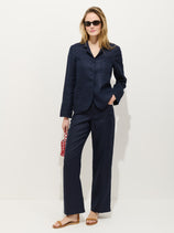 A woman in navy blue Lafayette Pant in linen with a matching blazer, wide-leg fit, tan sandals, sunglasses, and a small red patterned handbag stands against a white background.