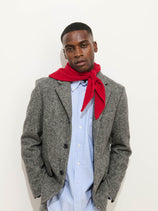 A man wearing a gray blazer, light blue shirt, and the Flynn Handkerchief Scarf in Wool in red stands against a plain white background with his hands in his pockets, looking at the camera.