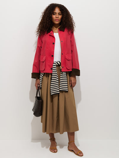 A woman stands against a white background wearing the Chiltern Cropped Jacket in waxed canvas, a white top, beige midi skirt, striped sweater tied at her waist, brown sandals, and holding a woven black handbag.