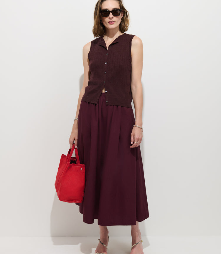 Woman in a sleeveless maroon top and matching Sarah Skirt In Cotton, wearing sunglasses, holding a bright red tote bag, and standing against a plain white background.