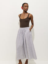 A person stands against a plain white background wearing the Sarah Striped Skirt in Linen—featuring thin blue vertical stripes—paired with a sleeveless brown tank top, hands in pockets and a red flower in their hair.