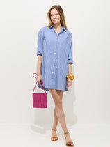A woman stands against a white background wearing the Florence Striped Shirt Dress in Poplin. She pairs it with tan sandals, a pink woven handbag, and a yellow scrunchie on her wrist.