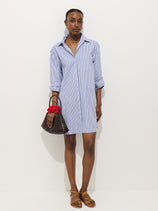 A woman stands against a white background wearing the Florence Striped Shirt Dress In Poplin. She holds a brown handbag with a red scrunchie, wears tan sandals, and has rolled-up sleeves for a relaxed fit.