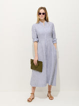 A woman in sunglasses wears the Isabella Striped Dress In Linen—a blue and white striped, 100% linen, long-sleeve midi shirt dress with rolled-up sleeves. She holds a green woven clutch and wears brown sandals against a plain white background.