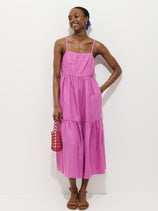 A person smiles while wearing the Clara Dress In Linen—an ankle-length, sleeveless pink dress with pockets and spaghetti straps. They hold a red woven handbag and wear brown sandals, posing against a plain white background.