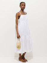 A woman models the Clara Dress In Linen, a sleeveless, white, ankle-length 100% linen dress. She stands against a plain white background, holding a small woven bag and wearing brown sandals.