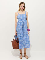 A woman wears the Clara Dress in Liberty Fabric, a blue floral sundress, with brown sandals, a woven brown tote bag, and a purple scrunchie on her wrist. Her hair is down and she stands against a plain background.