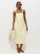 Wearing the Marie Dress In Linen—a pale yellow, 100% linen, midi bias cut—paired with brown sandals, sunglasses, and blue earrings, a person holds a beige and white striped sweater against a plain white background.