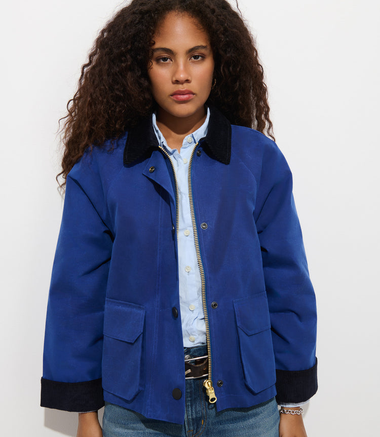 Wearing the Chiltern Cropped Jacket in Waxed Canvas in blue over a light blue shirt and jeans, a person with long curly hair stands against a plain white background with a neutral expression.