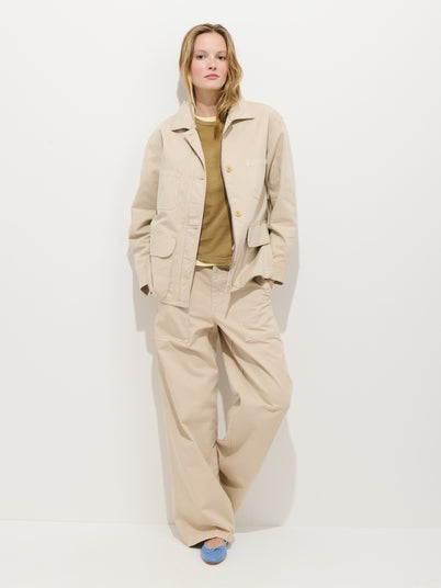 A person stands against a white wall in an oversized Painter Jacket In Herringbone and wide-leg trousers, with a tan shirt and light blue shoes. Their hands are in their pockets and they appear relaxed.