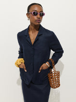 A stylish person wearing the Lafayette Jacket In Linen, a skirt, purple sunglasses, a yellow scrunchie on one wrist, and holding a brown woven handbag stands against a plain white background.
