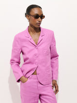 Wearing the Lafayette Jacket In Linen, a person poses against a white background with one hand in their pocket, looking off to the side and wearing sunglasses.