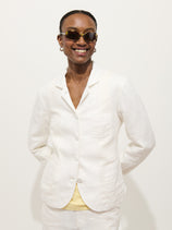 A smiling person wearing the Lafayette Jacket In Linen and sunglasses stands against a plain white background with their hands behind their back.