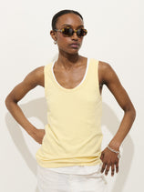Wearing the Garcon Tank In Linen Cotton over a white women’s tank, paired with white pants, sunglasses, hoop earrings, and a bracelet, a person stands with hands on hips against a plain background.