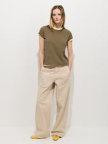 A person stands by a white wall, wearing the Prospect Tee in Cotton Jersey (olive green) and loose beige pants with large front pockets. With their hands in their pockets, shoulder-length hair, and yellow shoes, they complete this cotton look.