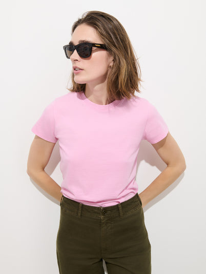 A woman wearing sunglasses and the Prospect Tee In Cotton Jersey—a light pink, 100% cotton, garment-dyed T-shirt made in Peru—paired with olive green pants stands against a plain white background with her hands behind her back, looking slightly to the side. The Prospect Tee offers both comfort and effortless style.