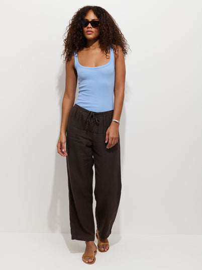 A woman with curly hair, black sunglasses, a light blue sleeveless top, sandals, bracelet, and wide-leg Hudson Pant In Linen with a pull-on waist stands against a plain white background.