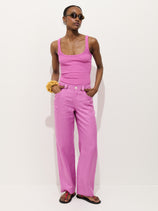 A person models the Lafayette Pant in Linen—100% linen, garment dyed—paired with a sleeveless pink tank top. They accessorize with sunglasses, a yellow scrunchie, and sandals, standing against a white background with hands in pockets.