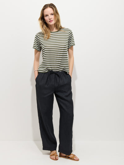 A person with medium-length blond hair stands against a white background wearing a green and white striped T-shirt, brown sandals, and the Hudson Pant In Linen with a pull-on waist. Their hands are in their pockets.